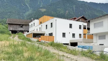 Modernes Gebäude mit weißer Fassade und Holzverkleidung vor alpiner Bergkulisse.