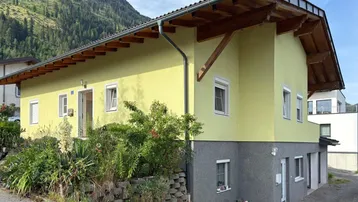 Hausfassade in Gelb mit Holzelementen und Bergblick, umgeben von grüner Natur.