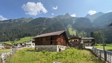 Altes Holzgebäude in ländlicher Umgebung mit grünen Wiesen und majestätischen Bergen im Hintergrund.