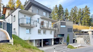 Modernes Mehrfamilienhaus mit Balkonen und Tiefgaragenzufahrt, umgeben von grüner Landschaft und Bäumen.