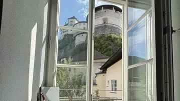 Helles Fenster mit Blick auf eine historische Burg auf einem grünen Hügel unter blauem Himmel.