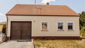 Traditionelles Einfamilienhaus mit beiger Fassade, braunem Garagentor und Ziegeldach unter blauem Himmel.