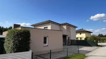 Modernes Einfamilienhaus mit beigefarbener Fassade und gepflasterter Einfahrt unter blauem Himmel.