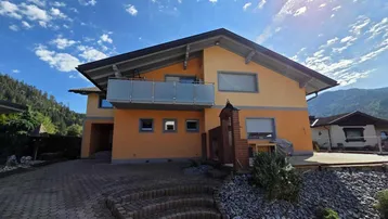 Vorderansicht des Hauses mit Balkon, gepflasterter Auffahrt und Bergblick an einem sonnigen Tag.