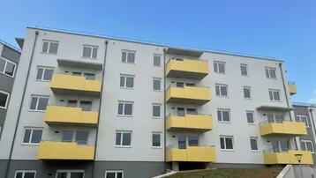 Modernes Mehrfamilienhaus mit hellgrauer Fassade und gelben Balkonen unter blauem Himmel.
