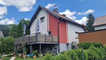 Modernes Einfamilienhaus mit roter Fassade, großem Balkon und Blick auf die umliegende Natur.