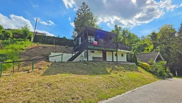 Freistehendes Haus mit dunkler Holzfassade und Balkon auf einem sonnigen Hanggrundstück.