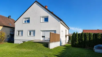 Weißes Einfamilienhaus mit Satteldach und großem Gartenbereich unter blauem Himmel.