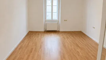 Helles, leeres Zimmer mit Parkettboden, weißen Wänden und einem klassischen Fenster.