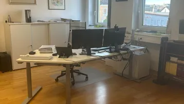 Geräumiges Büro mit zwei Fenstern, Holzfußboden und modernem Schreibtisch mit Computern.