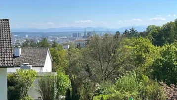 Weitblick über grüne Baumwipfel auf die Stadt und die dahinterliegende Bergkette unter blauem Himmel.