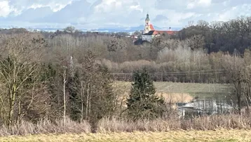 Fernblick auf eine Kirche und die umliegende Landschaft mit Feldern und Bäumen unter bewölktem Himmel.