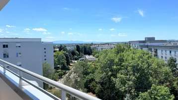 Weitblick vom Balkon über die Stadt und grüne Umgebung bis zu den fernen Bergen.