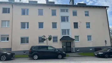 Mehrfamilienhaus mit beiger Fassade und geparkten Autos davor unter bewölktem Himmel.
