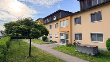 Mehrfamilienhaus mit gepflegter Fassade, Grünflächen und Bäumen unter blauem Himmel.