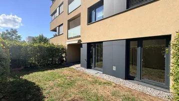 Kleiner Gartenbereich vor einem modernen Gebäude mit großen Fenstern und viel Sonnenlicht.