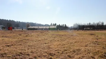 Weitläufiges Feld mit Blick auf entfernte Gebäude und bewaldete Hügel unter blauem Himmel.