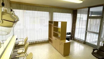 Geräumiger Innenraum eines Friseursalons mit mehreren Arbeitsplätzen, großen Fenstern und Jalousien, gut beleuchtet.