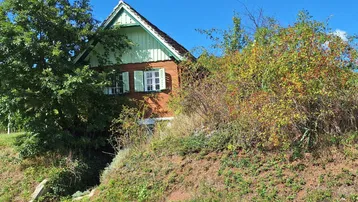Kleines Haus mit grünen Fensterläden und Holzfassade, umgeben von üppiger Vegetation auf einem Hang.