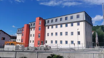 Neubau-Gebäude mit moderner Fassade und roten Akzenten unter blauem Himmel.