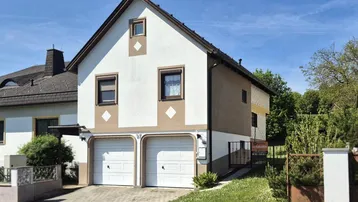 Vorderansicht des Hauses mit zwei Garagen und gepflegtem Vorgarten unter blauem Himmel.