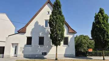 Weißes Einfamilienhaus mit Giebeldach und einem hohen Baum davor, an einer ruhigen Straße gelegen.
