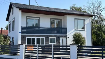 Modernes Einfamilienhaus mit weißer Fassade, dunklem Dach und Balkon, umgeben von einem Zaun.