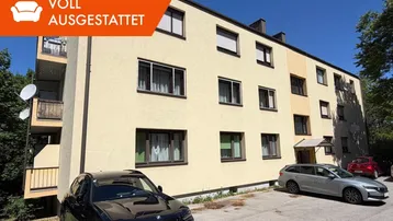 Mehrfamilienhaus mit mehreren Balkonen und ausreichend Parkplätzen im Freien, umgeben von Grünflächen.