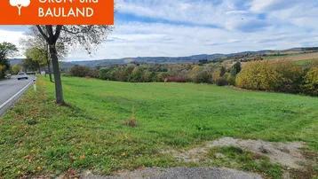 Großes grünes Feld neben einer Straße mit Hügeln und Bäumen im Hintergrund unter blauem Himmel.