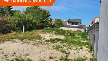 Unbebautes Grundstück mit leichter Hanglage, umgeben von Vegetation und Wohnhäusern unter blauem Himmel.