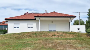 Modernes Einfamilienhaus mit weißer Fassade, rotem Ziegeldach und Rollläden, umgeben von einer grünen Wiese.