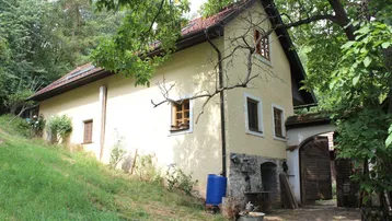 Traditionelles Landhaus mit Putzfassade und Holztüren, umgeben von viel Grün und Bäumen.