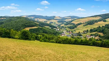 Weitläufige Landschaft mit grünen Hügeln, Wäldern und einem kleinen Dorf im Tal unter blauem Himmel.