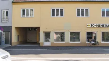 Gebäudefassade mit gelber Farbe, rotem Ziegeldach und mehreren Fenstern. Im Erdgeschoss befinden sich Ladenflächen und eine Garageneinfahrt.