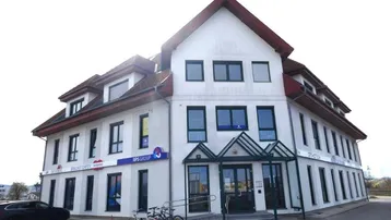 Gebäude mit weißer Fassade, rotem Ziegeldach und mehreren Fenstern, inklusive barrierefreiem Eingangsbereich mit Rampe.