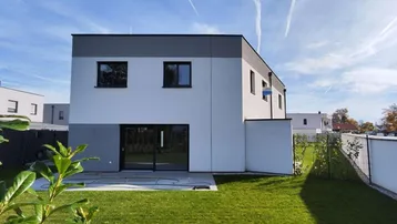 Moderne Hausfassade mit großen Glasfronten und gepflegtem Rasenbereich unter blauem Himmel.