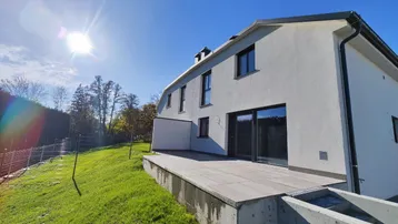 Moderne Hausfassade mit großer gepflasterter Terrasse und sonnigem Ausblick ins Grüne.