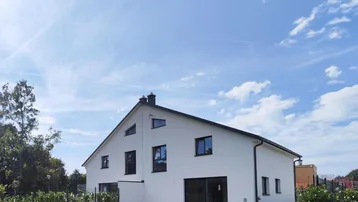 Modernes Einfamilienhaus mit weißer Fassade, schwarzem Dach und gepflegtem Rasen unter blauem Himmel.