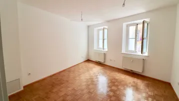 Helles Zimmer mit Parkettboden, weißen Wänden und zwei Fenstern, die viel Tageslicht hereinlassen.