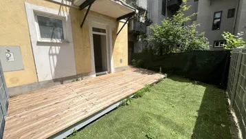 Ebenerdige Holzterrasse mit Zugang zum Gebäude, kleinem Rasen und Balkon darüber.
