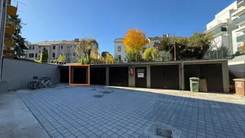 Reihe von Garagenboxen auf einem gepflasterten Parkplatz unter blauem Himmel mit Bäumen im Hintergrund.