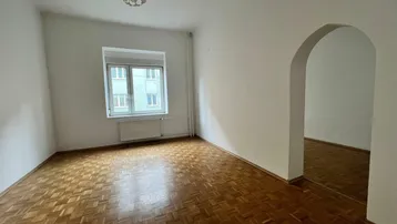 Helles Zimmer mit klassischem Parkettboden, großem Fenster und einem eleganten Bogen zum Nebenraum.
