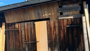 Alte Holzscheune mit verwitterten Holztüren und Brettern unter einem Baum, die als Lager dient.