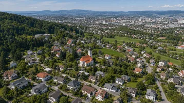 Luftaufnahme einer Wohnsiedlung mit Kirche, umgeben von viel Grün und einer Stadt im Hintergrund.