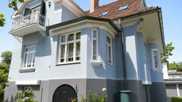 Historisches Haus mit hellblauer Fassade, roten Ziegeln und kleinem Balkon.