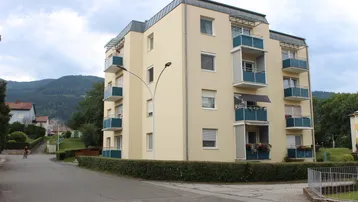 Mehrstöckiges Wohngebäude mit beiger Fassade und blauen Balkonen in einer grünen, bergigen Umgebung.