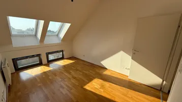 Helles Zimmer im Dachgeschoss mit Holzboden, schrägen Wänden und mehreren Dachfenstern für viel Tageslicht.