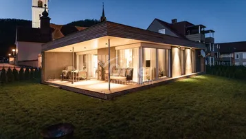 Moderne Architektur mit großer Holzterrasse und gemütlicher Sitzecke, beleuchtet bei Dämmerung.