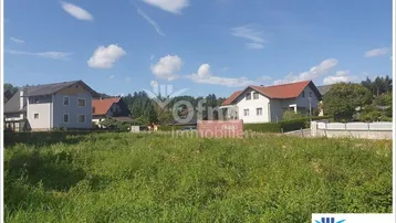 Ungenutztes Grundstück mit grüner Wiese und Wohnhäusern im Hintergrund unter blauem Himmel.