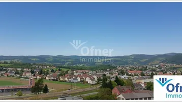 Weitläufiger Panoramablick über eine ländliche Siedlung mit grünen Hügeln im Hintergrund unter blauem Himmel.